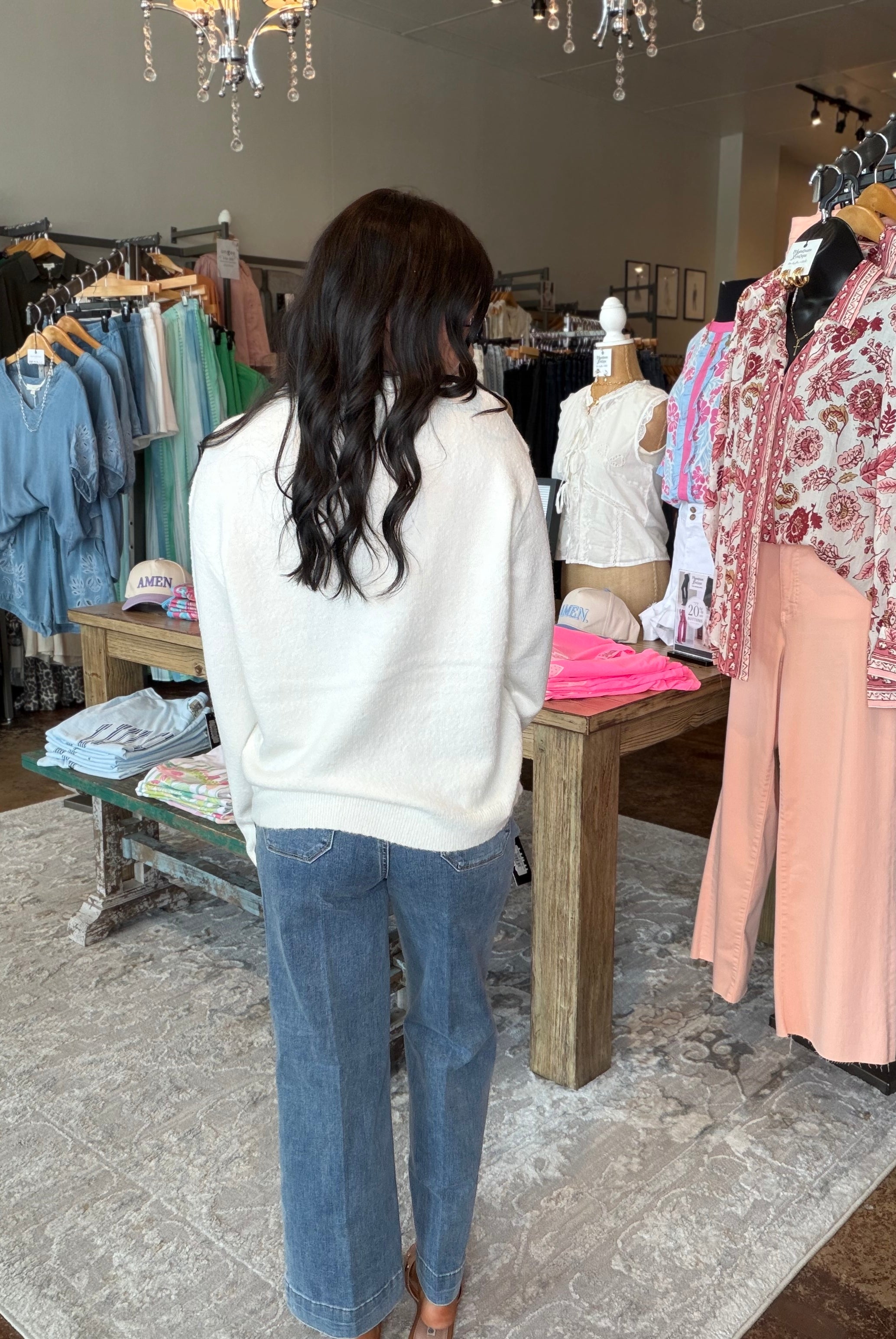 A woman with long dark hair stands with her back to the camera inside a clothing boutique, wearing a cream knit sweater and blue wide-leg jeans. Clothing racks with various garments are visible on both sides of the store.