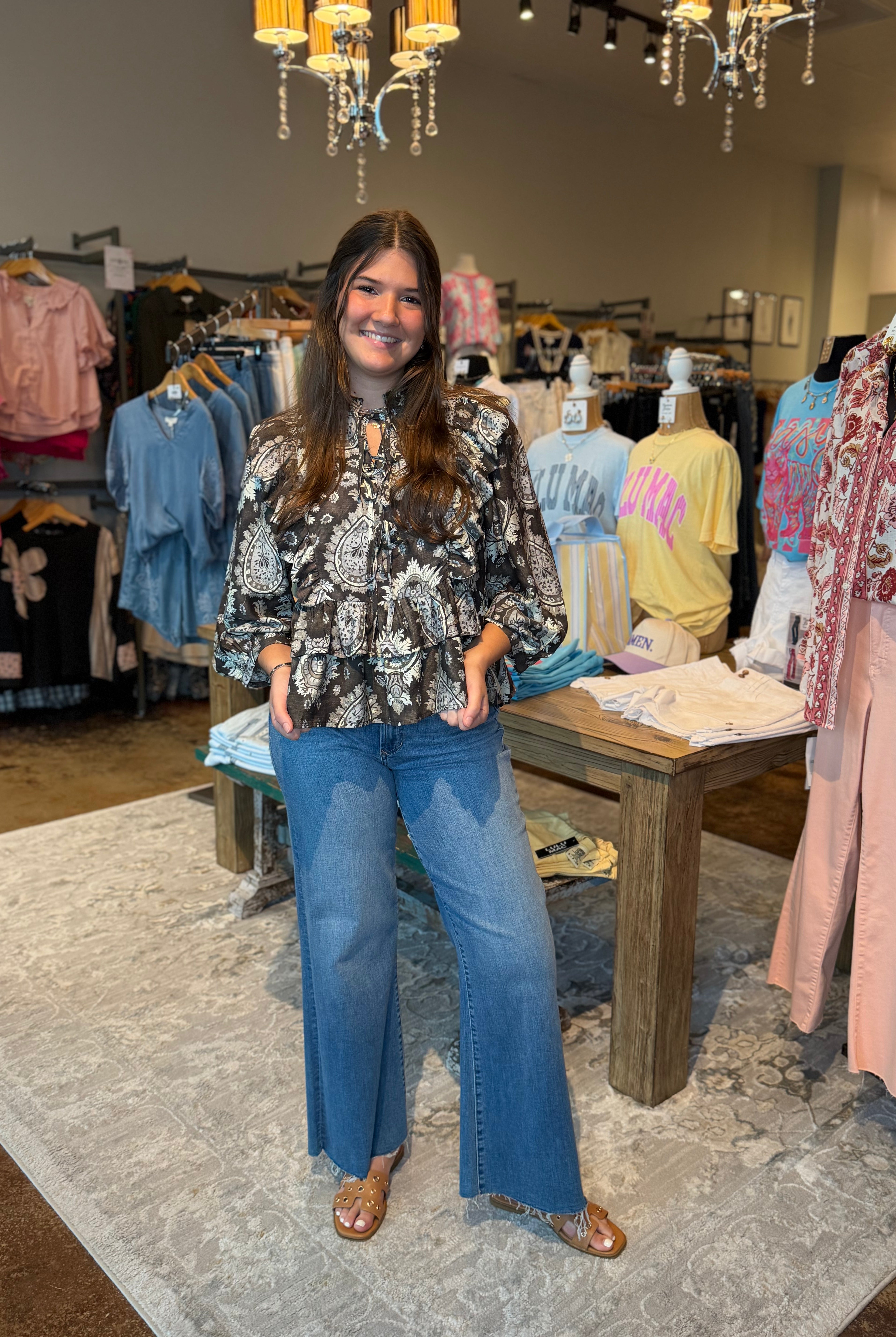 A woman stands in a clothing boutique wearing a black and white paisley patterned long-sleeve blouse with blue wide-leg jeans and brown flat sandals. The store has clothing racks, mannequins, a wooden table, and hanging chandeliers in the background.