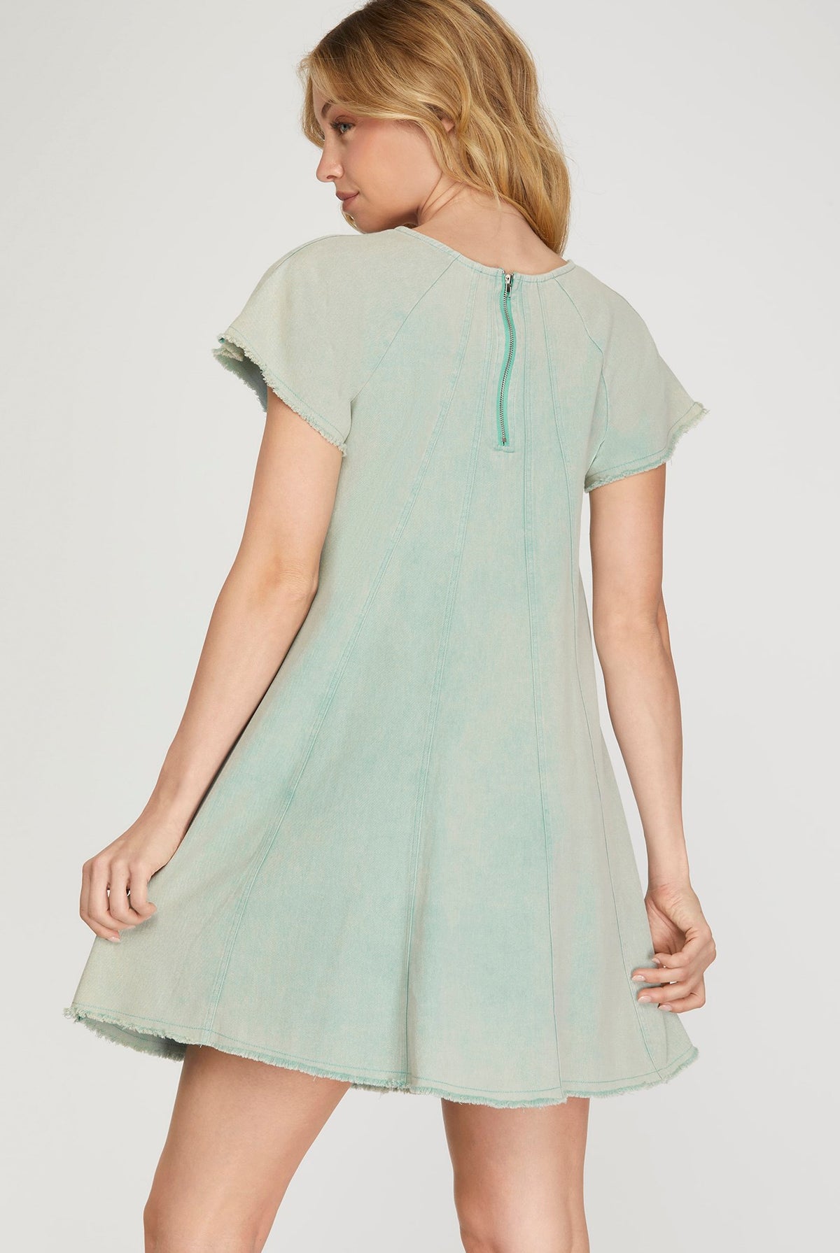 A woman wearing a light sage green short-sleeve dress, shown from the back against a plain light gray studio background.