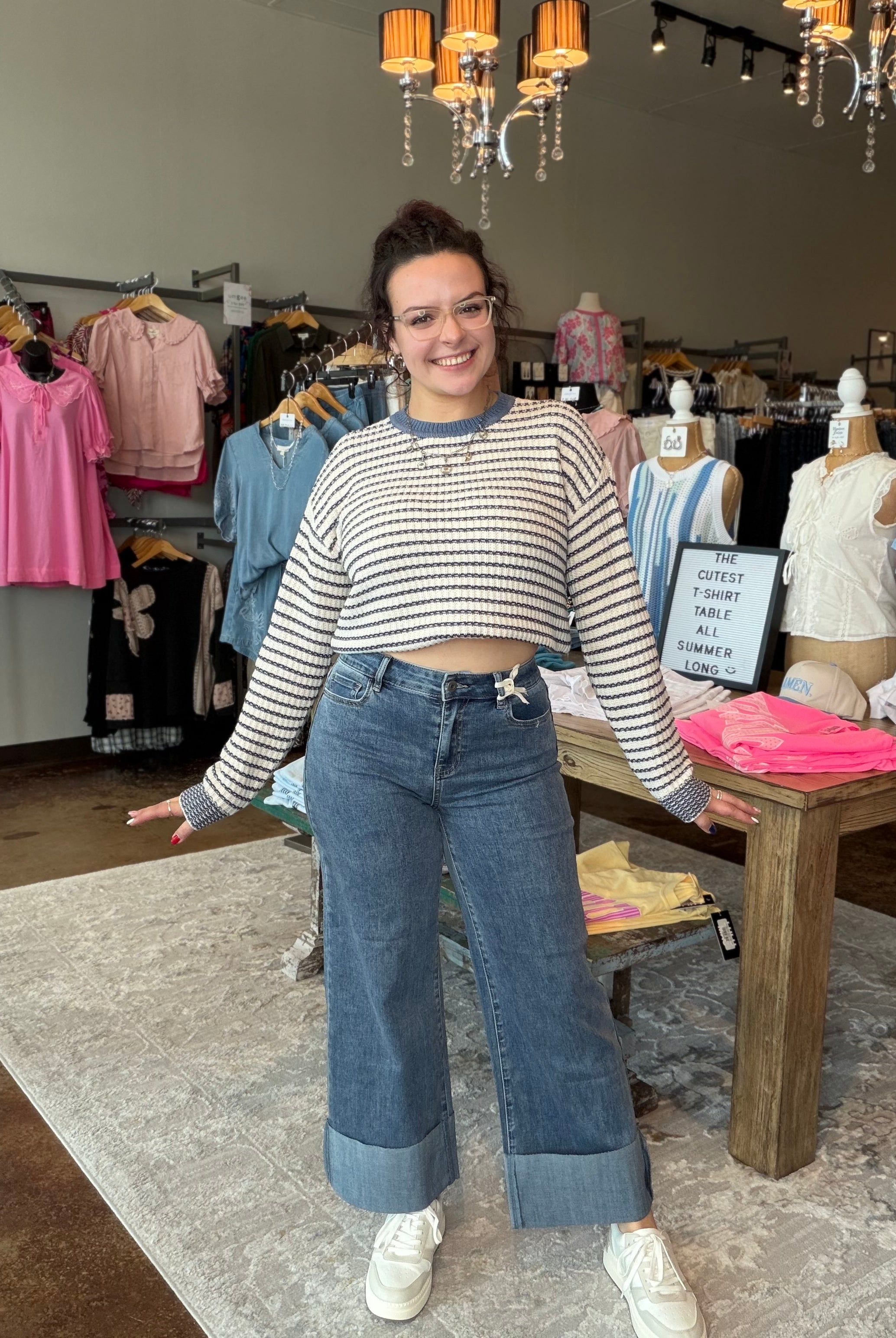 A woman modeling a striped long-sleeve crop top and wide-leg blue jeans with rolled cuffs, standing inside a clothing boutique.