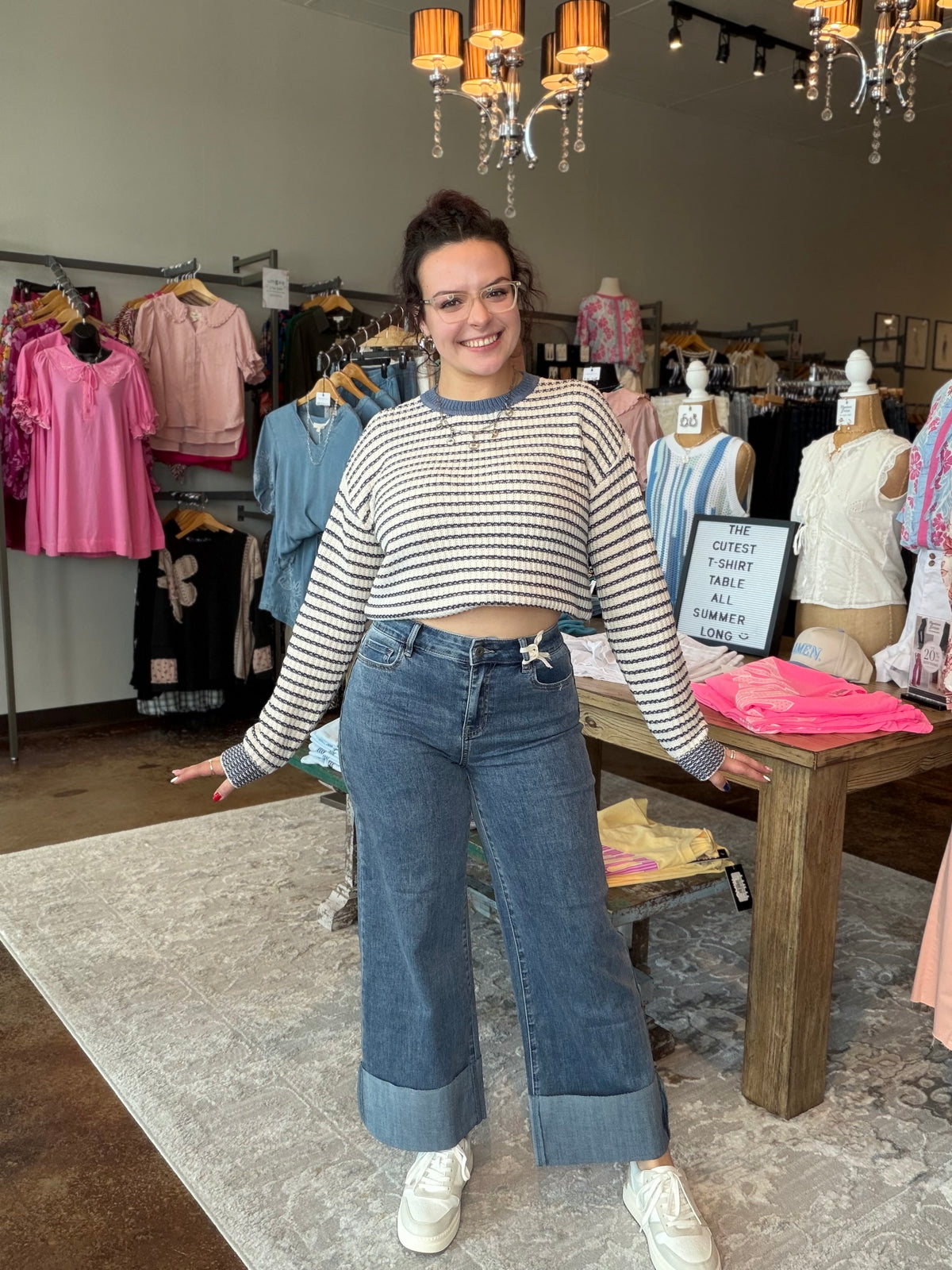 A woman modeling a striped long-sleeve crop top and wide-leg blue jeans with rolled cuffs, standing inside a clothing boutique.