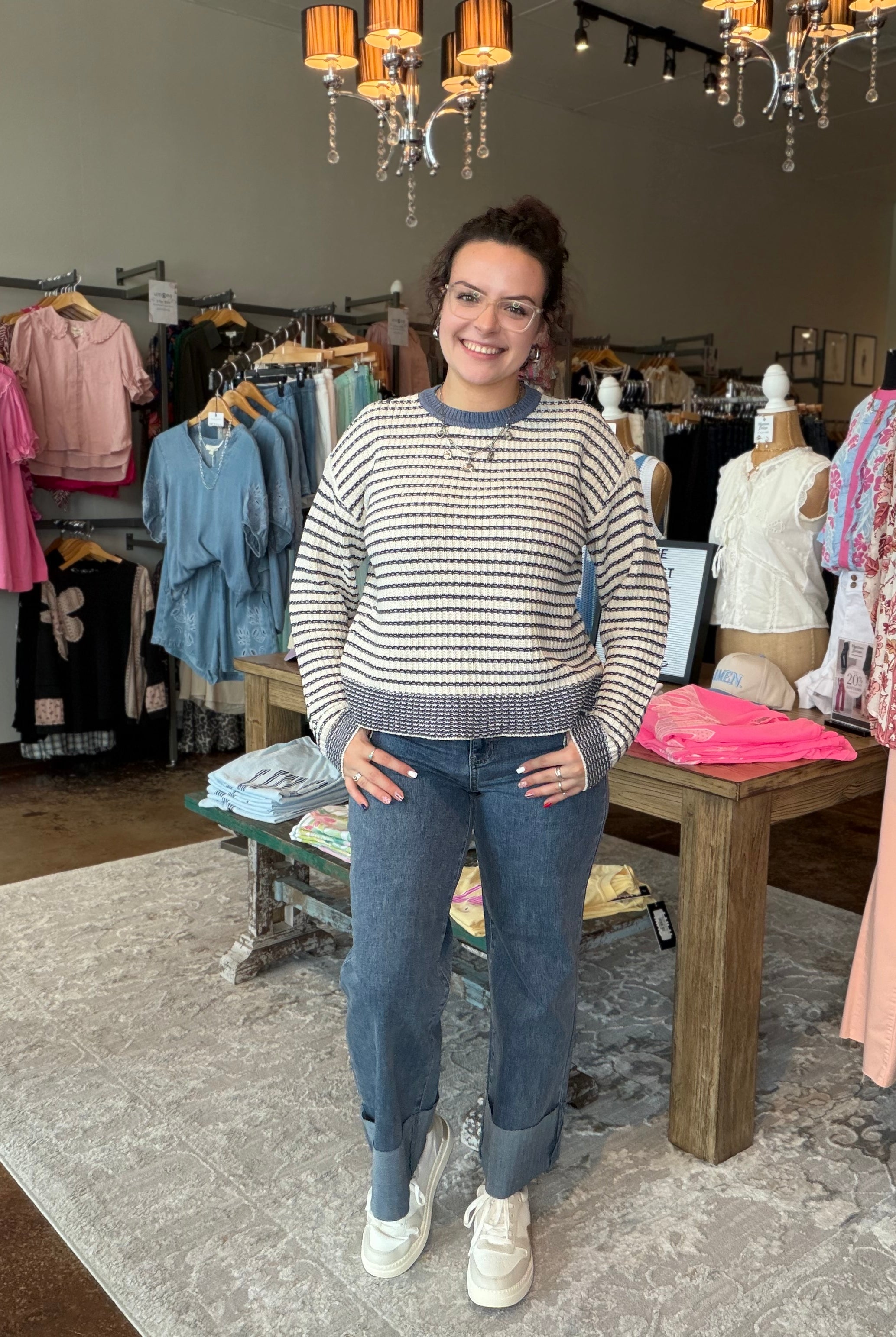 A woman stands in a clothing boutique wearing a cream and navy blue horizontally striped knit sweater, blue jeans, and white sneakers. The store is filled with racks of colorful clothing, mannequins, and hanging lights in the background.