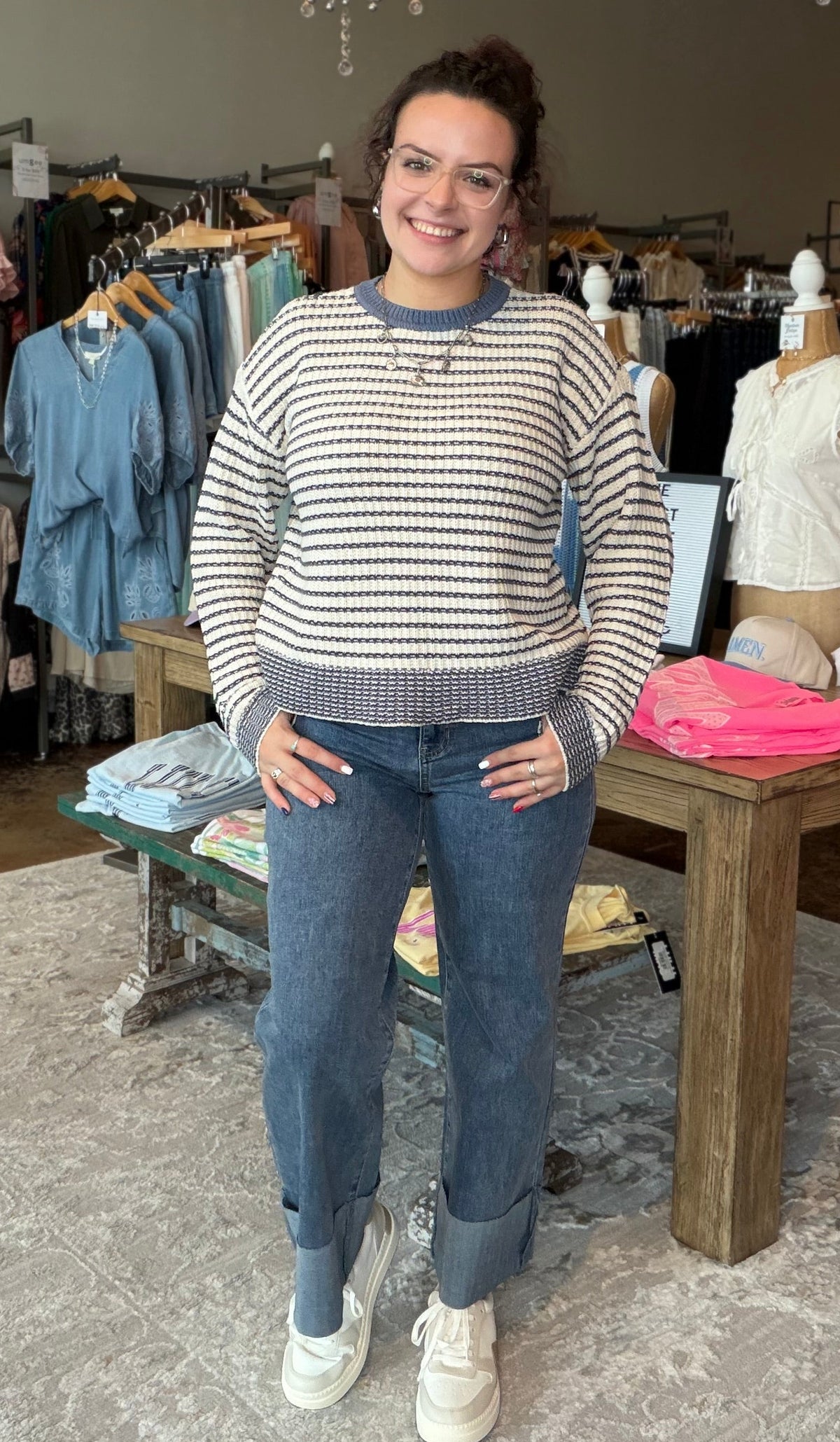 A woman stands in a clothing boutique wearing a cream and navy blue horizontally striped knit sweater, blue jeans, and white sneakers. The store is filled with racks of colorful clothing, mannequins, and hanging lights in the background.