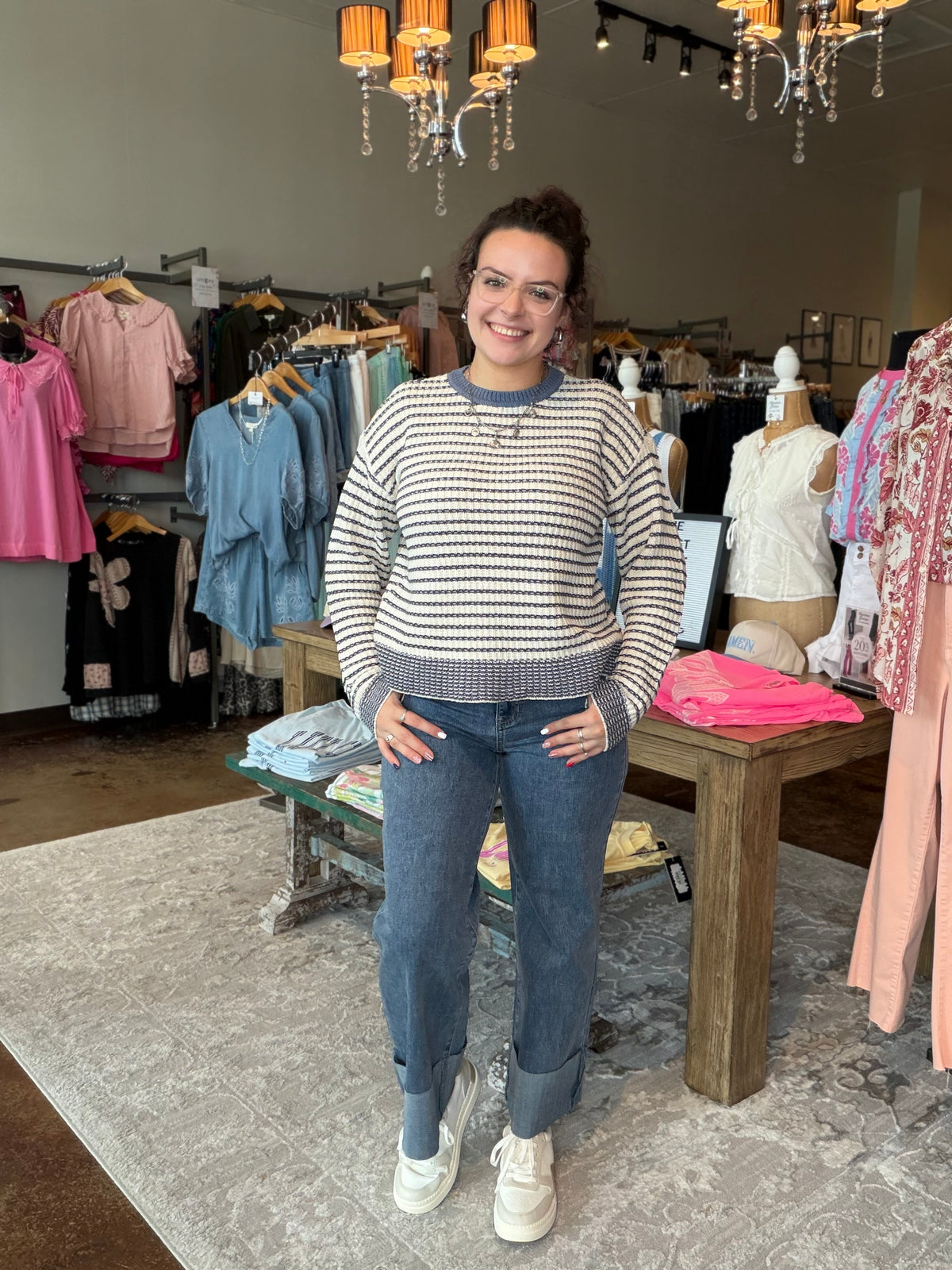 A woman stands in a clothing boutique wearing a cream and navy blue horizontally striped knit sweater, blue jeans, and white sneakers. The store is filled with racks of colorful clothing, mannequins, and hanging lights in the background.