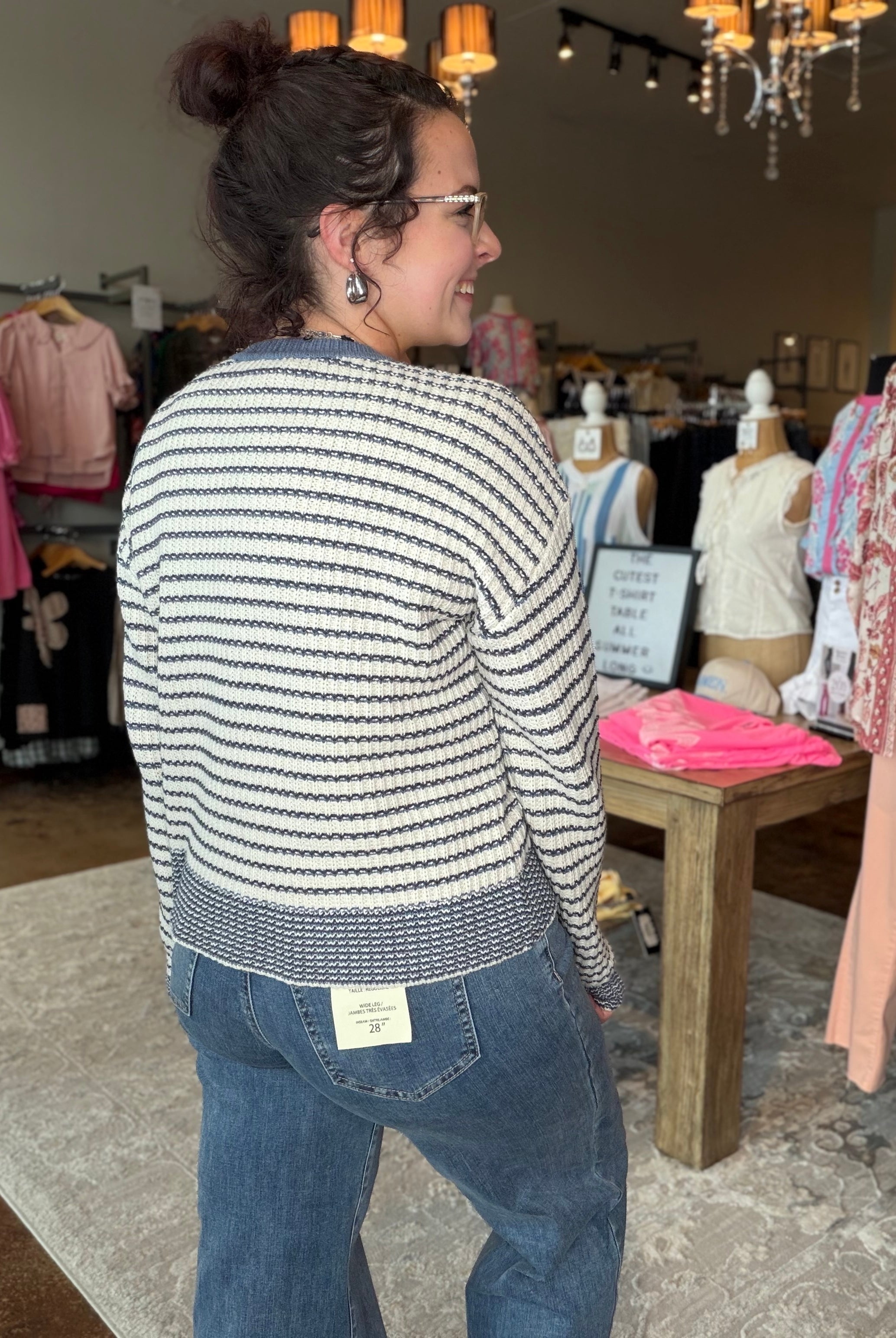 A woman wearing a cream and navy blue horizontally striped knit sweater and blue jeans, standing inside a clothing boutique.