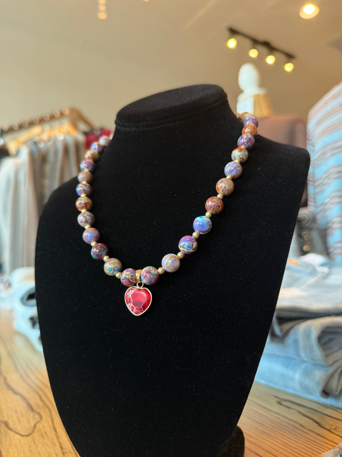 A beaded necklace with marbled purple, blue, and pink beads and a red heart-shaped pendant, displayed on a black velvet jewelry bust in a boutique setting.