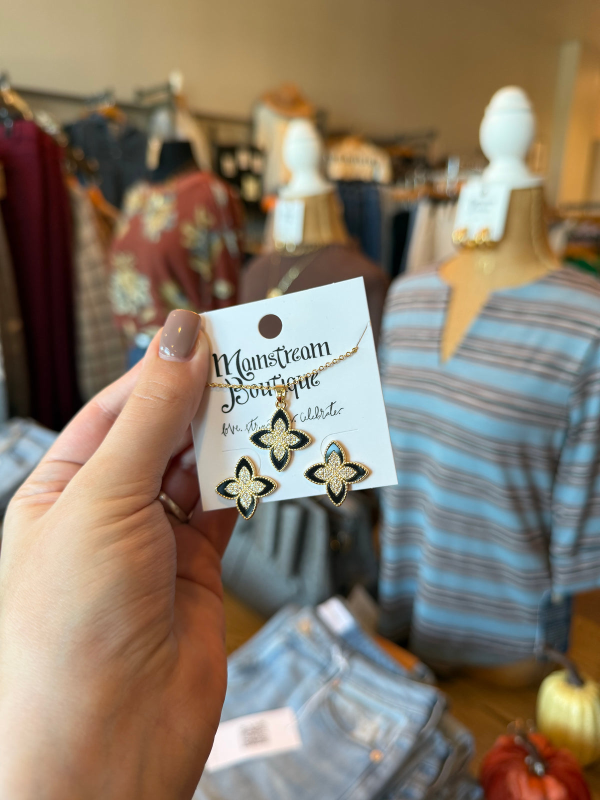 A hand holds a card displaying a pair of gold and black butterfly earrings in a boutique setting with clothing mannequins in the background.