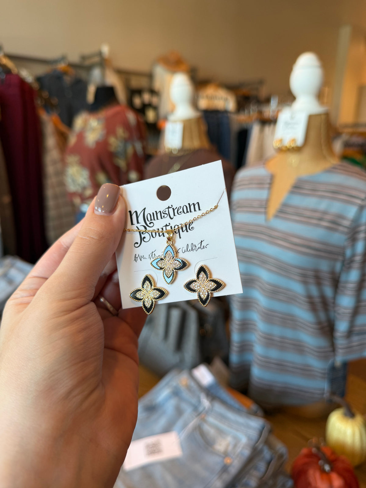 A hand holds a jewelry card displaying a gold-tone floral earring and matching necklace with turquoise and white accents, set against a blurred boutique background with mannequins and clothing racks.