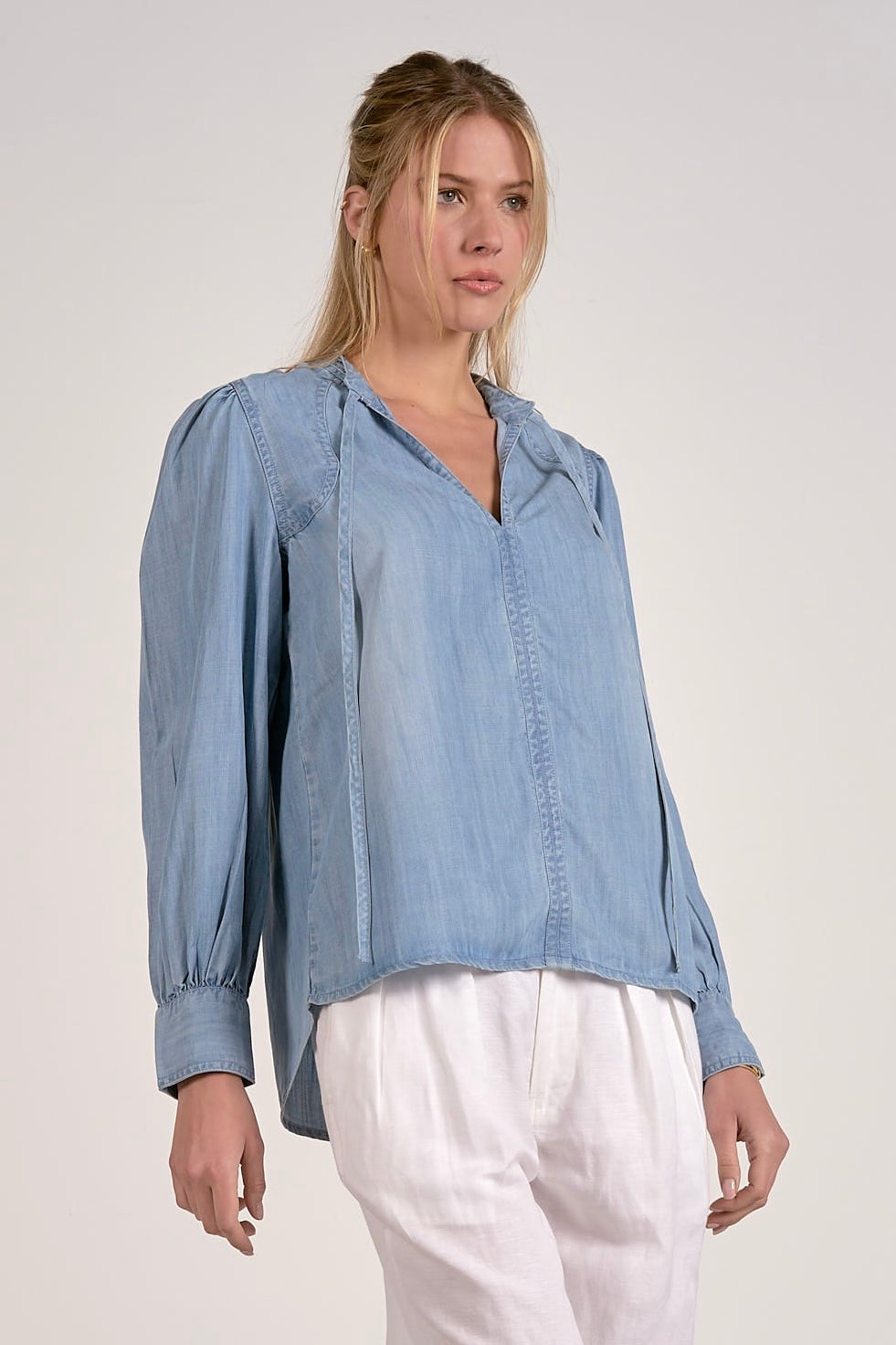 A woman wearing a light blue long-sleeve button-up blouse with gathered shoulders and white pants, photographed against a plain light gray studio background.