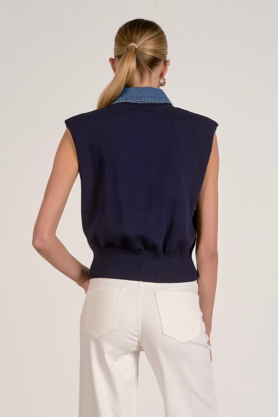 A woman shown from the back wearing a navy sleeveless knit vest over a blue denim collared shirt, paired with white trousers, against a light gray studio background.