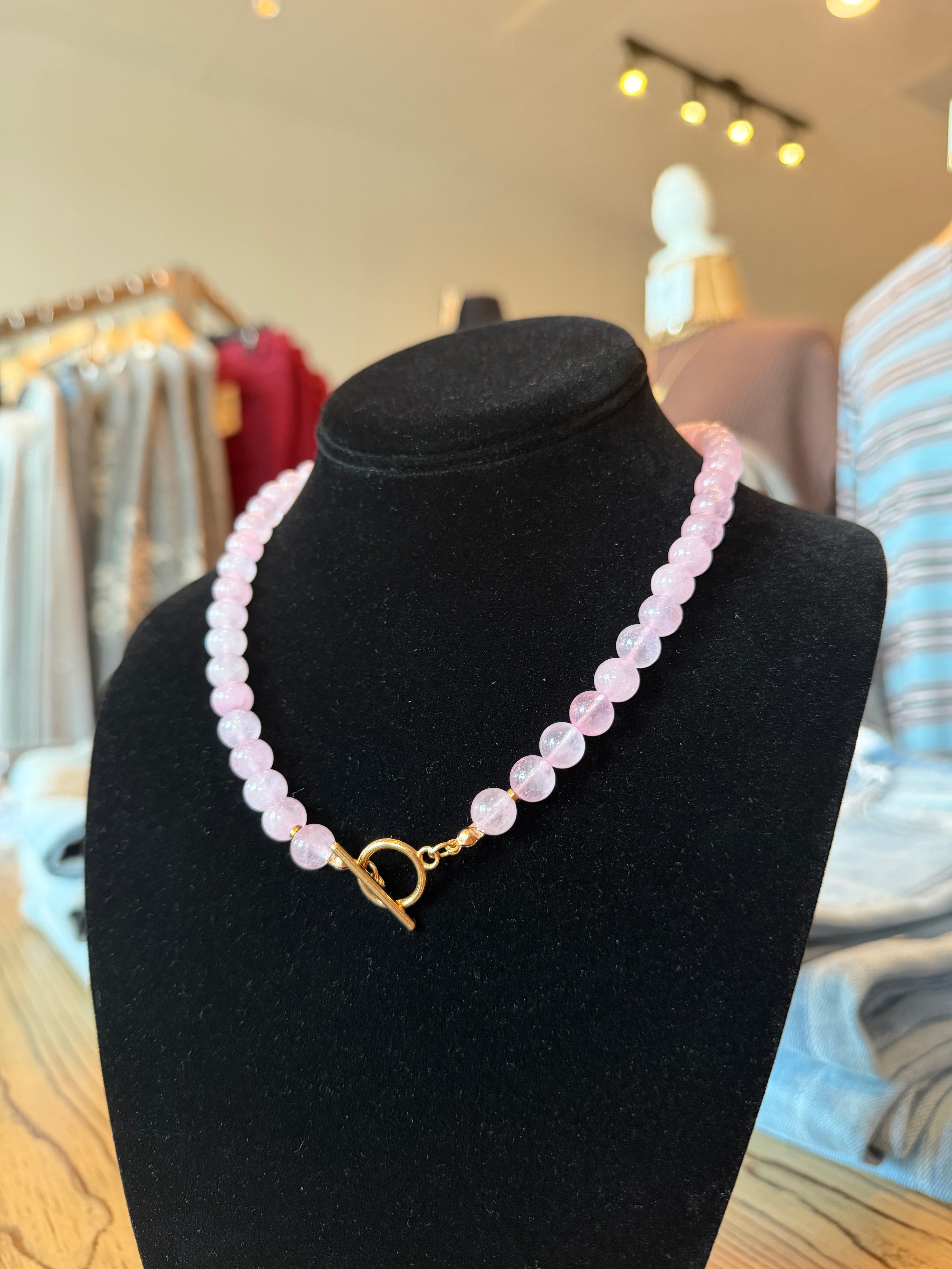 A pink beaded necklace with a gold toggle clasp displayed on a black velvet jewelry bust in a retail store setting.