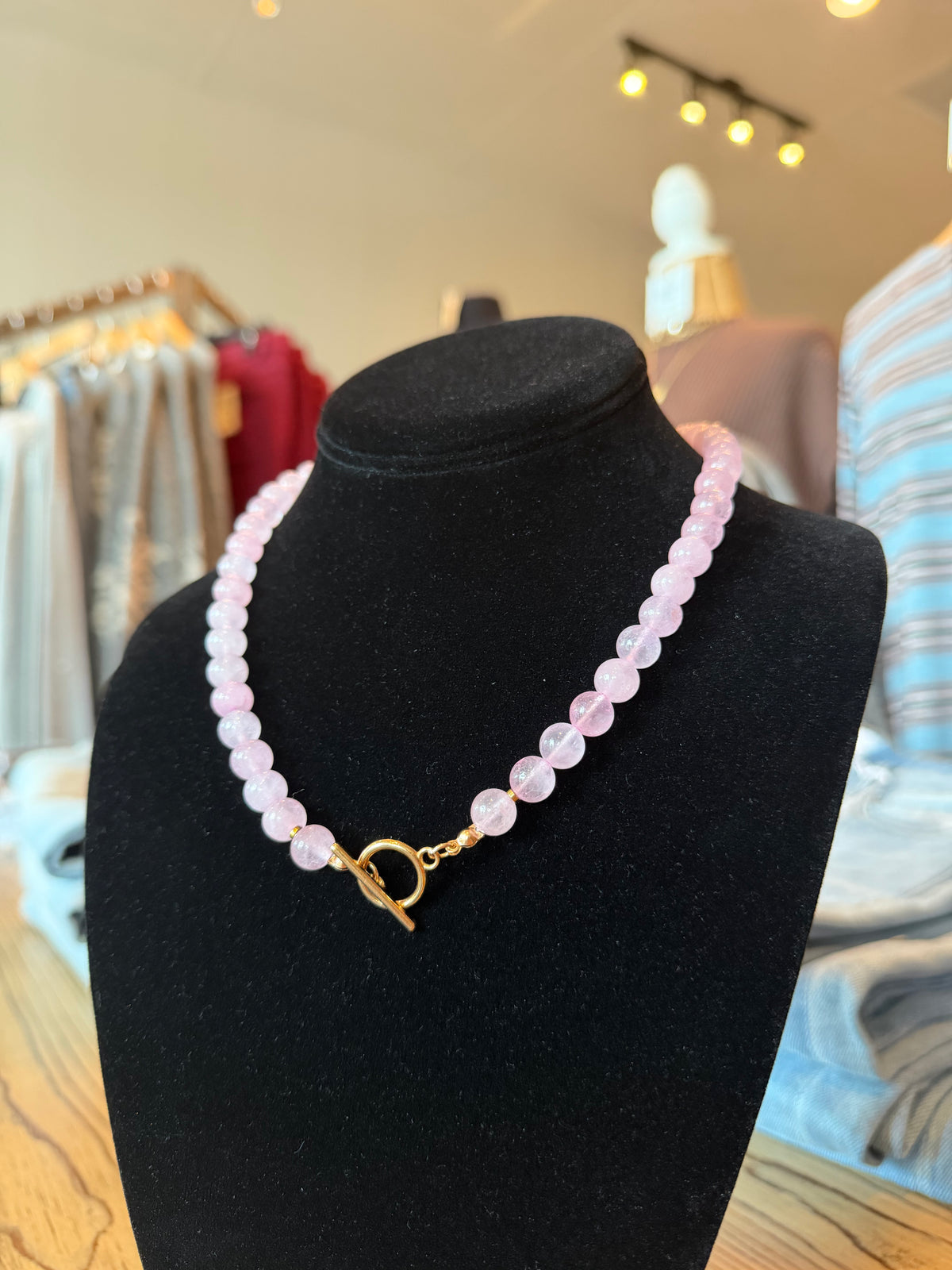 A pink beaded necklace with a gold toggle clasp displayed on a black velvet jewelry bust in a retail store setting.