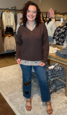 A woman stands in a clothing boutique wearing a brown v-neck sweater, a striped shirt, and blue jeans with heart patches. Clothing racks, a table with folded garments, and a patterned rug are visible in the background.