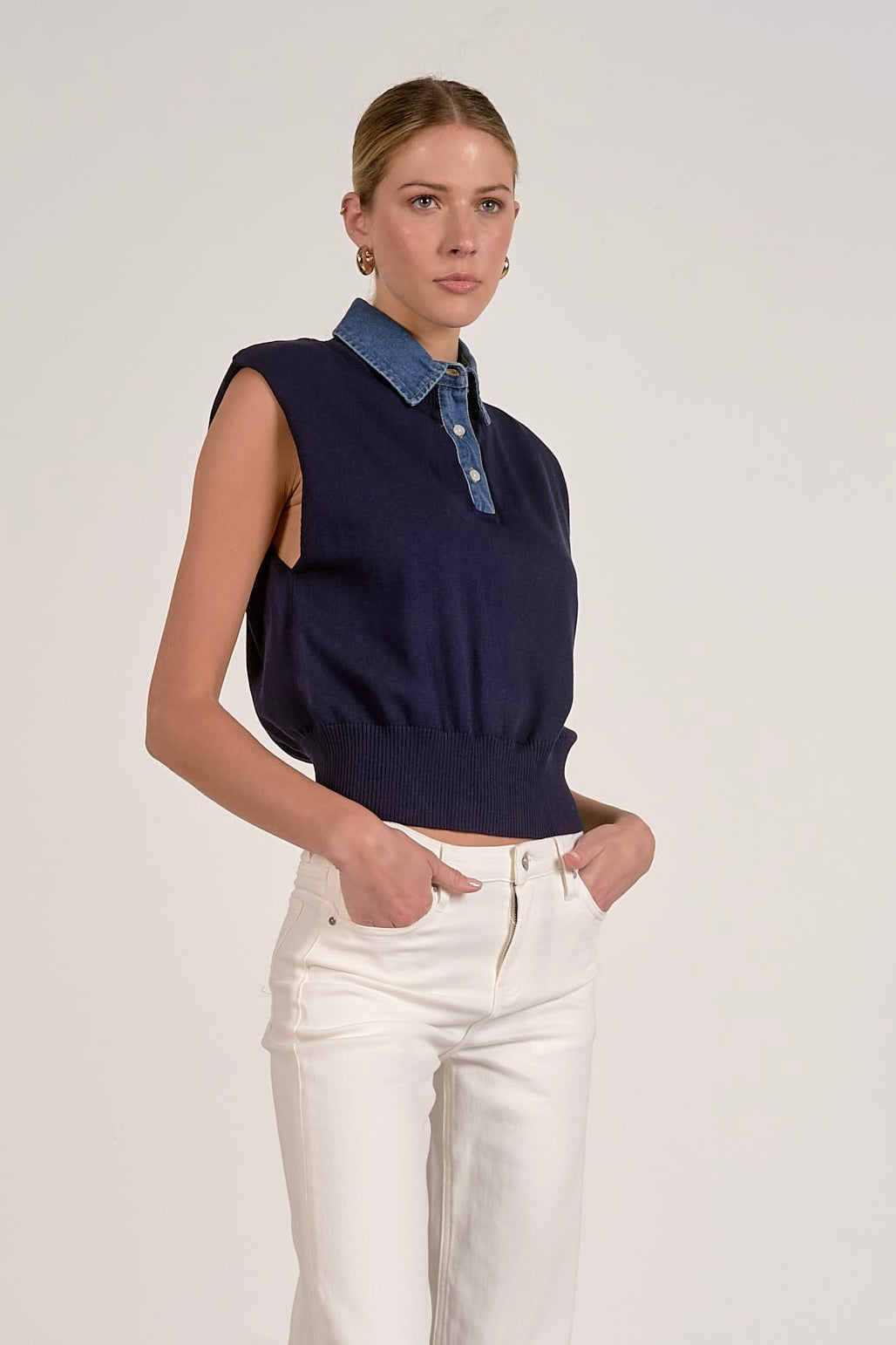A woman modeling a sleeveless navy knit top with a blue denim collar and white pants against a plain light gray studio background.