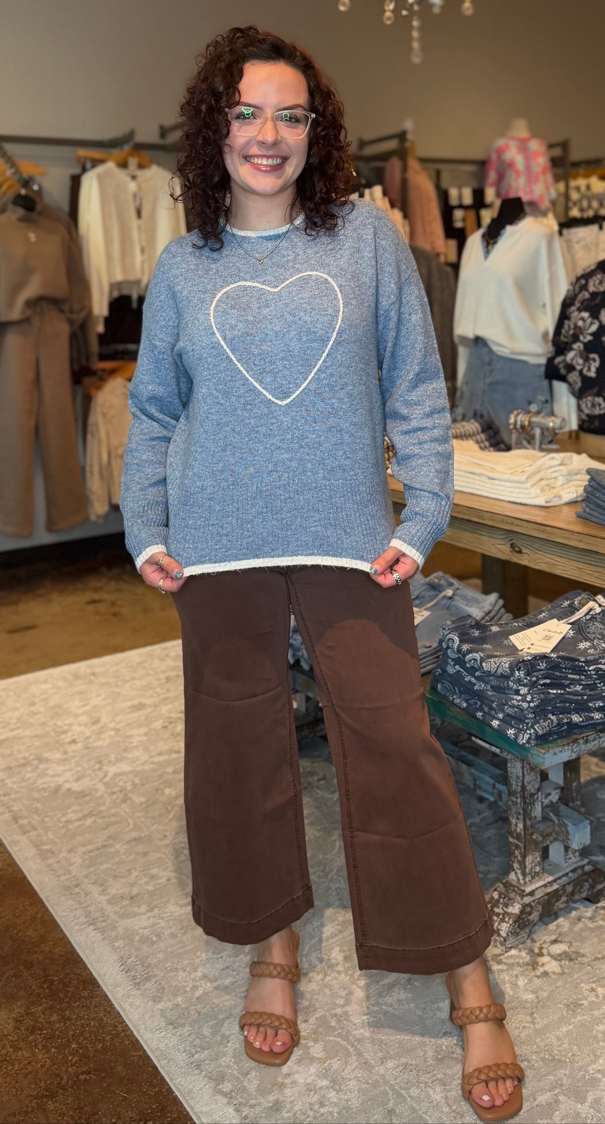 A woman stands in a clothing boutique wearing a light blue crewneck sweater with a white heart outline on the front, paired with brown wide-leg pants and tan sandals.