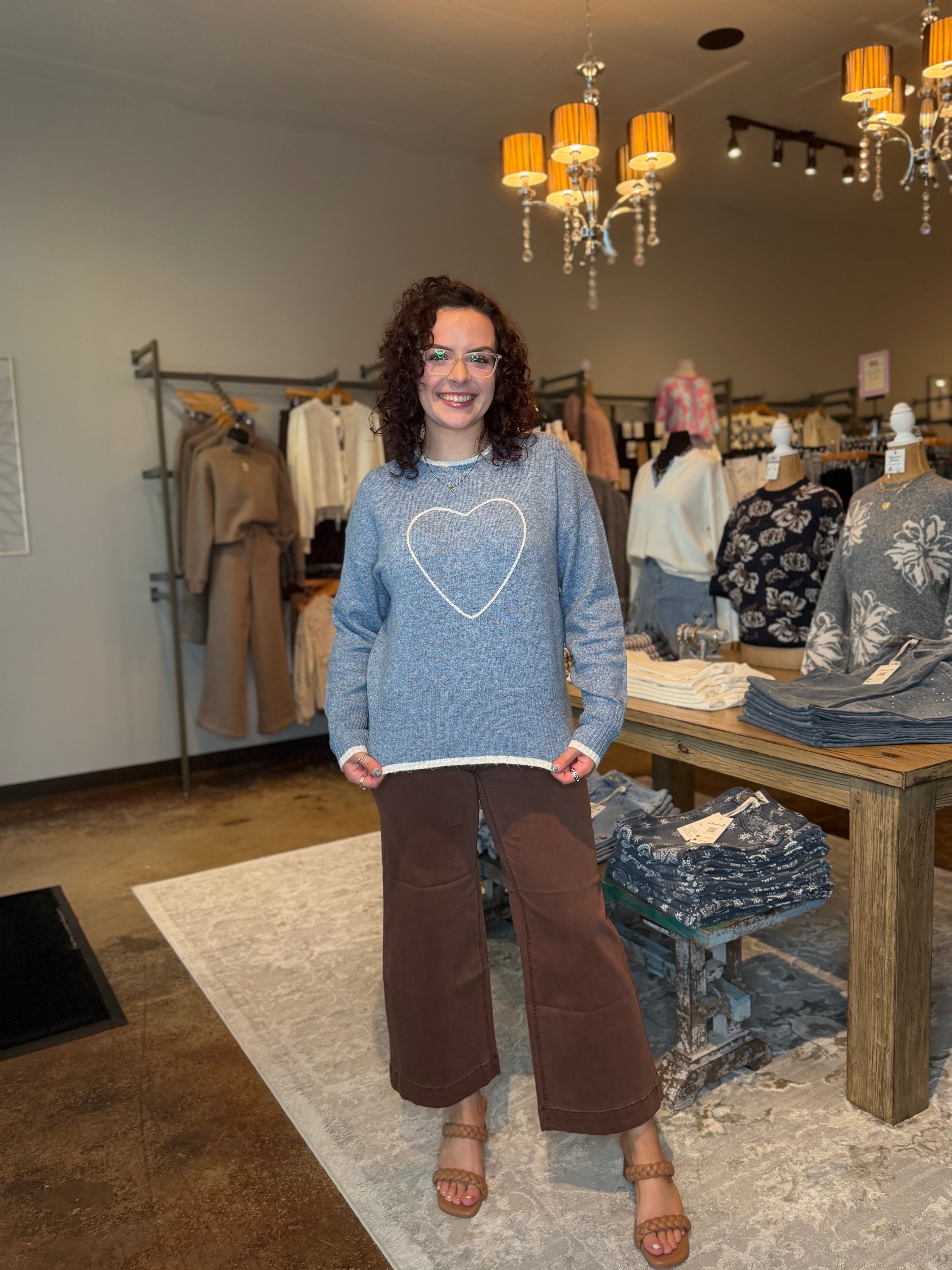 A woman stands in a clothing boutique wearing a light blue crewneck sweater with a white heart outline on the front, paired with brown wide-leg pants and tan sandals.