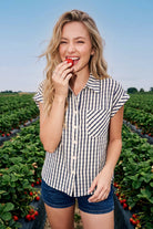 A woman standing in a strawberry field wearing a navy and white vertically striped short-sleeve button-up shirt and denim shorts, holding a strawberry to her mouth.