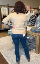 A woman stands with her back to the camera in a clothing boutique, wearing a beige knit sweater with blue floral patterned sleeves and blue jeans.