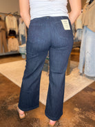 A person shown from the back wearing dark blue flare jeans in a clothing store. The jeans have a visible white brand label on the back pocket and are paired with a white top and beige sandals.
