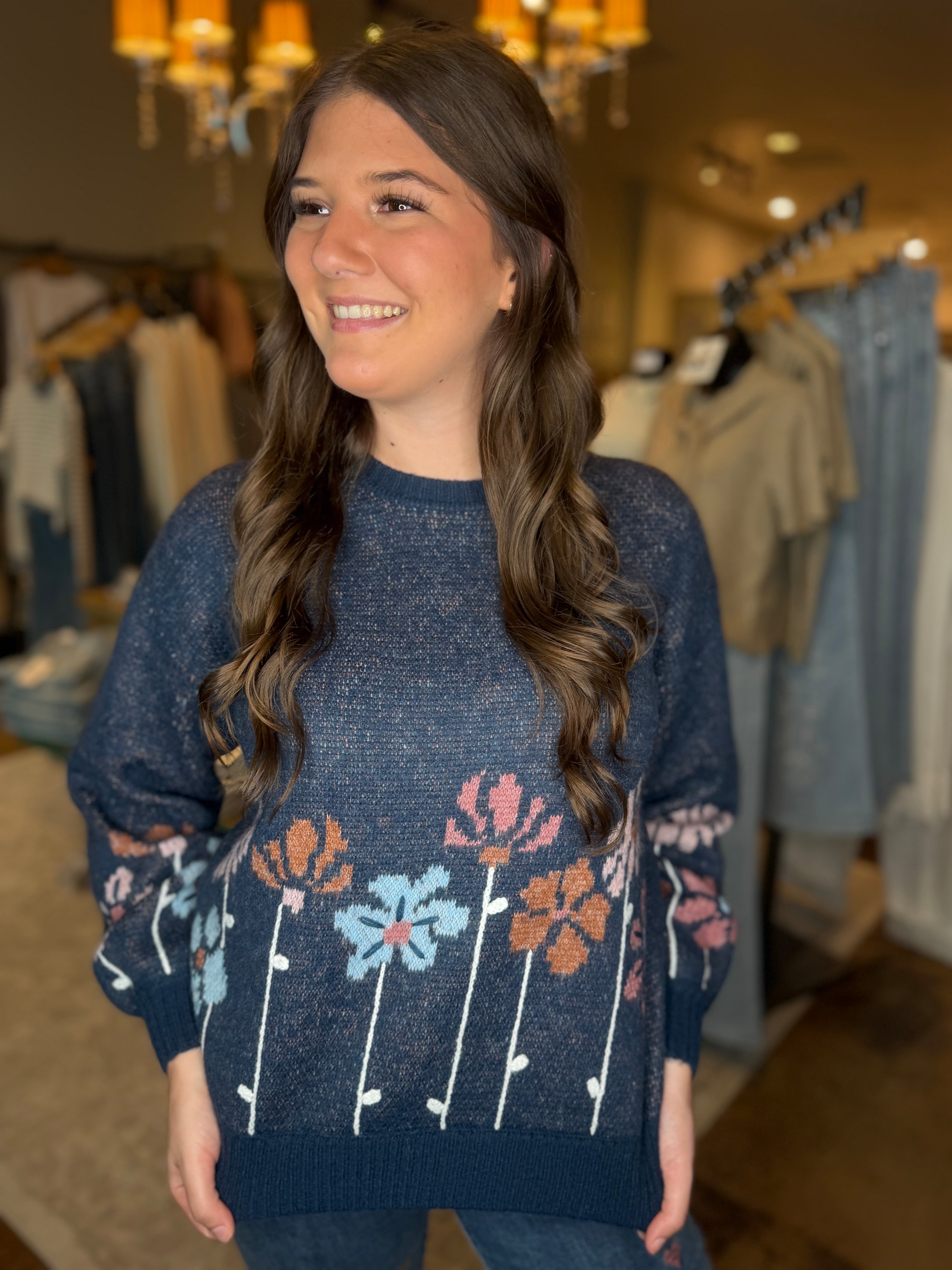 A woman with long brown hair smiling in a clothing store, wearing a navy blue knit sweater decorated with colorful floral patterns in pink, orange, and light blue.