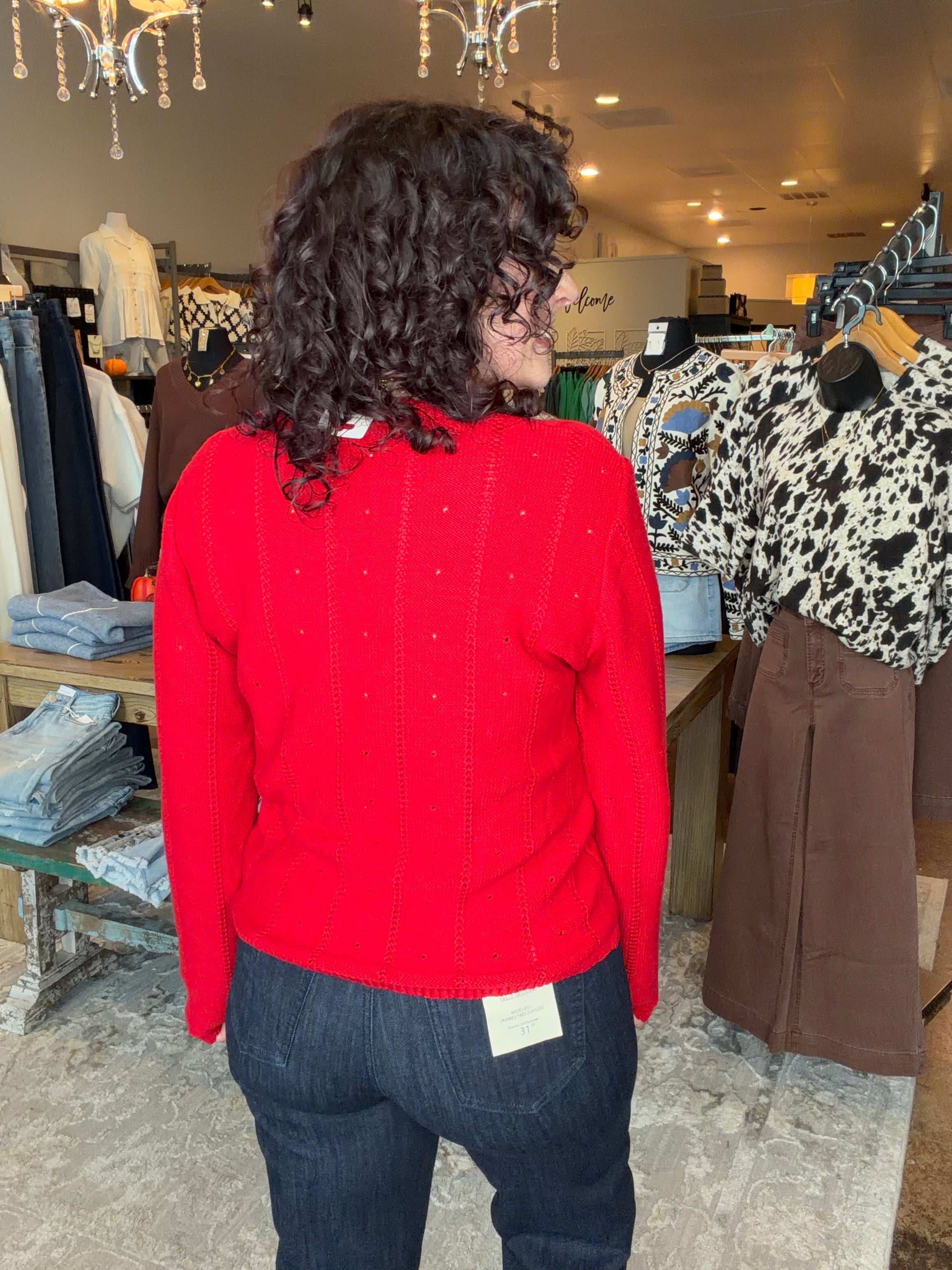 A person with curly dark hair seen from behind wearing a bright red knit sweater and dark blue jeans inside a clothing boutique. Other garments are visible on racks in the background.