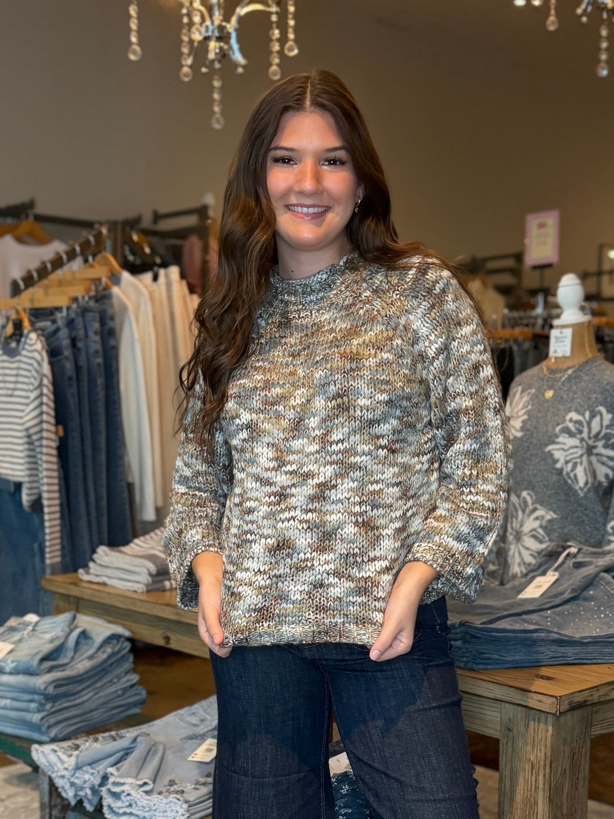A woman stands in a clothing boutique wearing a textured, long-sleeve knit sweater with a beige, brown, and gray abstract pattern, paired with dark blue wide-leg jeans and tan ankle boots.