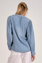 A woman shown from the back wearing a light blue long-sleeve blouse with gathered puff sleeves and white pants, photographed against a plain light background.