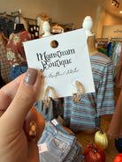 A hand holds a white display card featuring gold hoop earrings with clear stones, branded 'Mainstream Boutique'. The background shows a clothing boutique interior with mannequins, racks, and autumn decor.