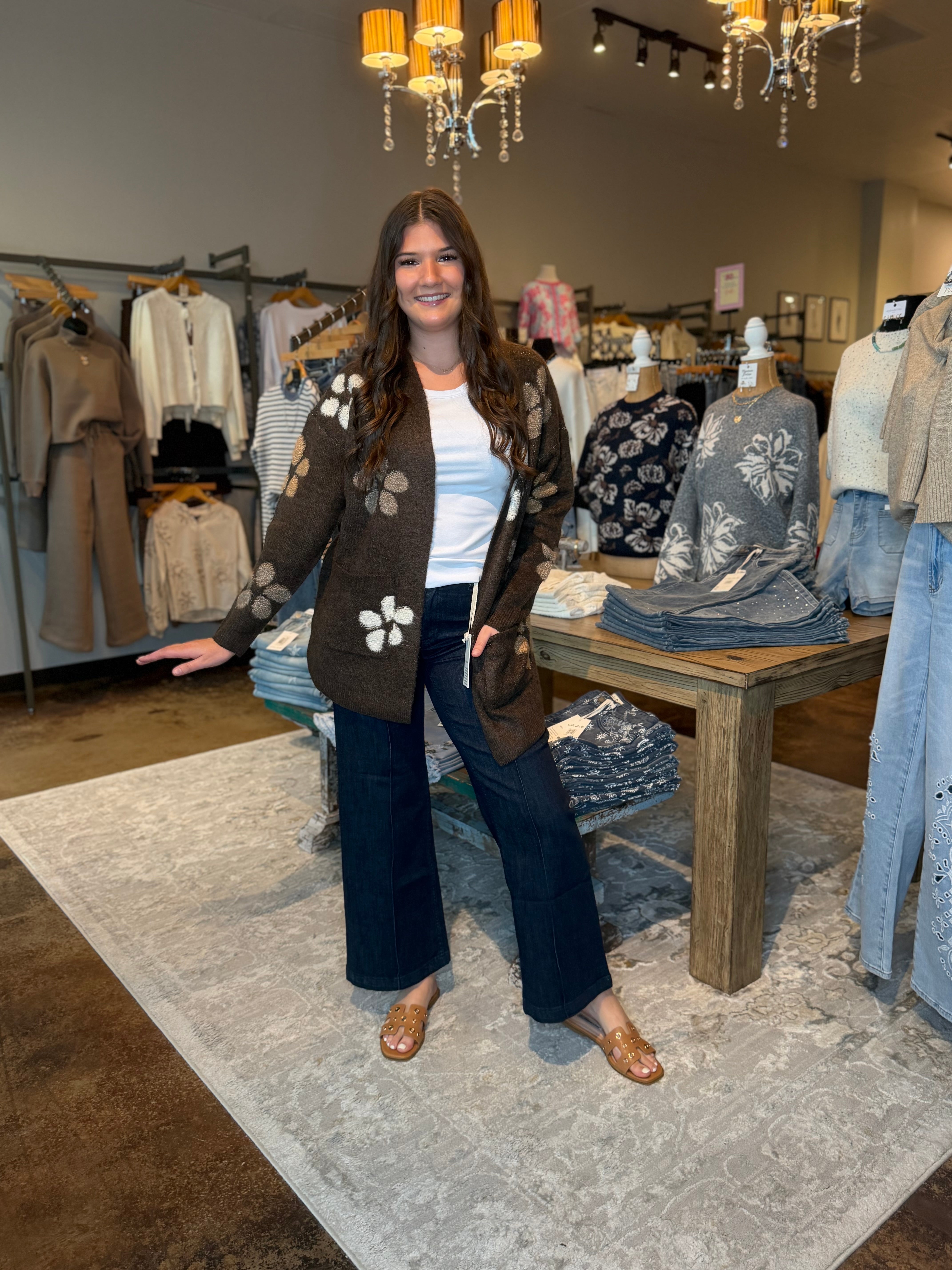 A woman stands in a boutique wearing a brown knit cardigan with white floral motifs, a white top, dark wide-leg pants, and brown flat shoes. The background shows clothing racks, mannequins, a wooden table with folded garments, and hanging lights.