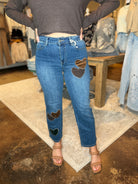 A person wearing blue jeans with brown heart patches on the front and lower legs, paired with a dark gray long-sleeve crop top and tan heeled sandals, standing in a clothing store.