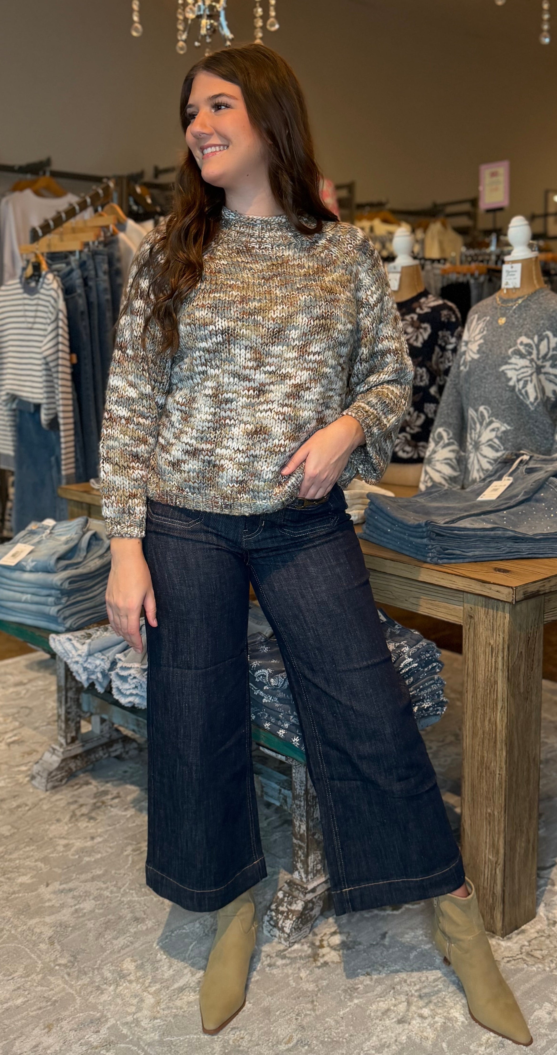 A woman stands in a clothing boutique wearing a textured knit sweater, dark wide-leg jeans, and tan ankle boots. The store has hanging garments, a wooden table with folded clothes, and warm hanging light fixtures in the background.