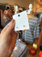 A hand holds a white display card featuring two small gold and white stud earrings with clear round backs, set against a busy boutique background with mannequins and clothing racks.
