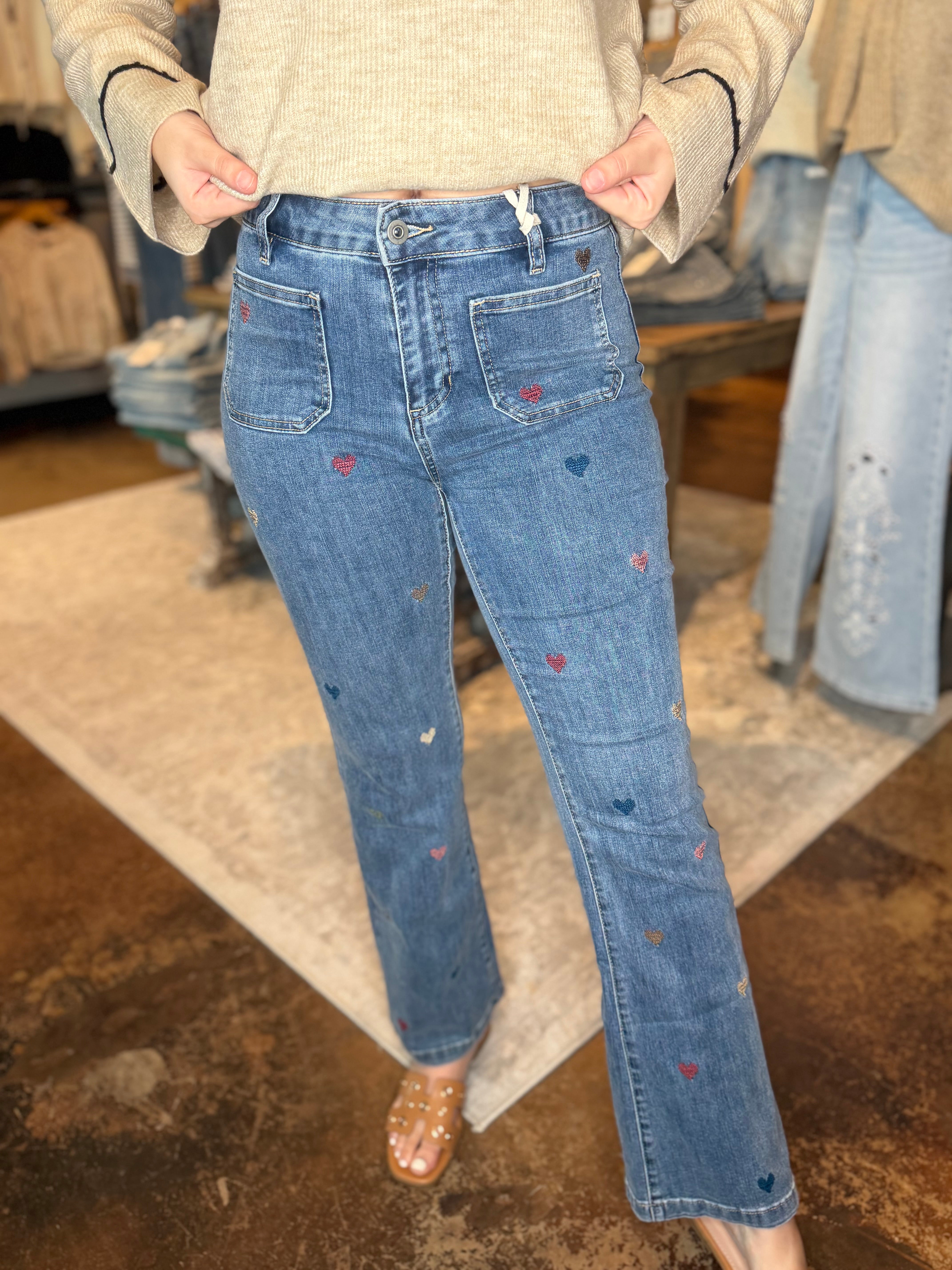 A person wearing blue denim jeans with small embroidered heart details in red, blue, and white, paired with a beige knit sweater and clear flat sandals in a retail store setting.