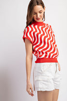 Woman wearing a red and white patterned top with white shorts on a light gray background
