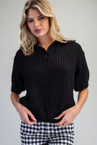 A woman wearing a black ribbed knit short-sleeve sweater with a split V-neck collar, paired with black and white gingham pants, against a plain light gray background.