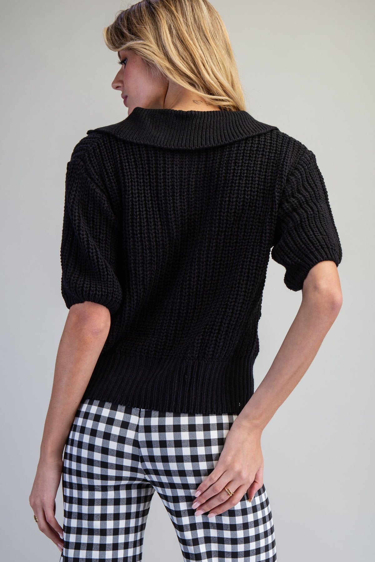 A woman shown from the back wearing a black short-sleeve ribbed knit sweater and black-and-white gingham pants against a plain light gray studio background.