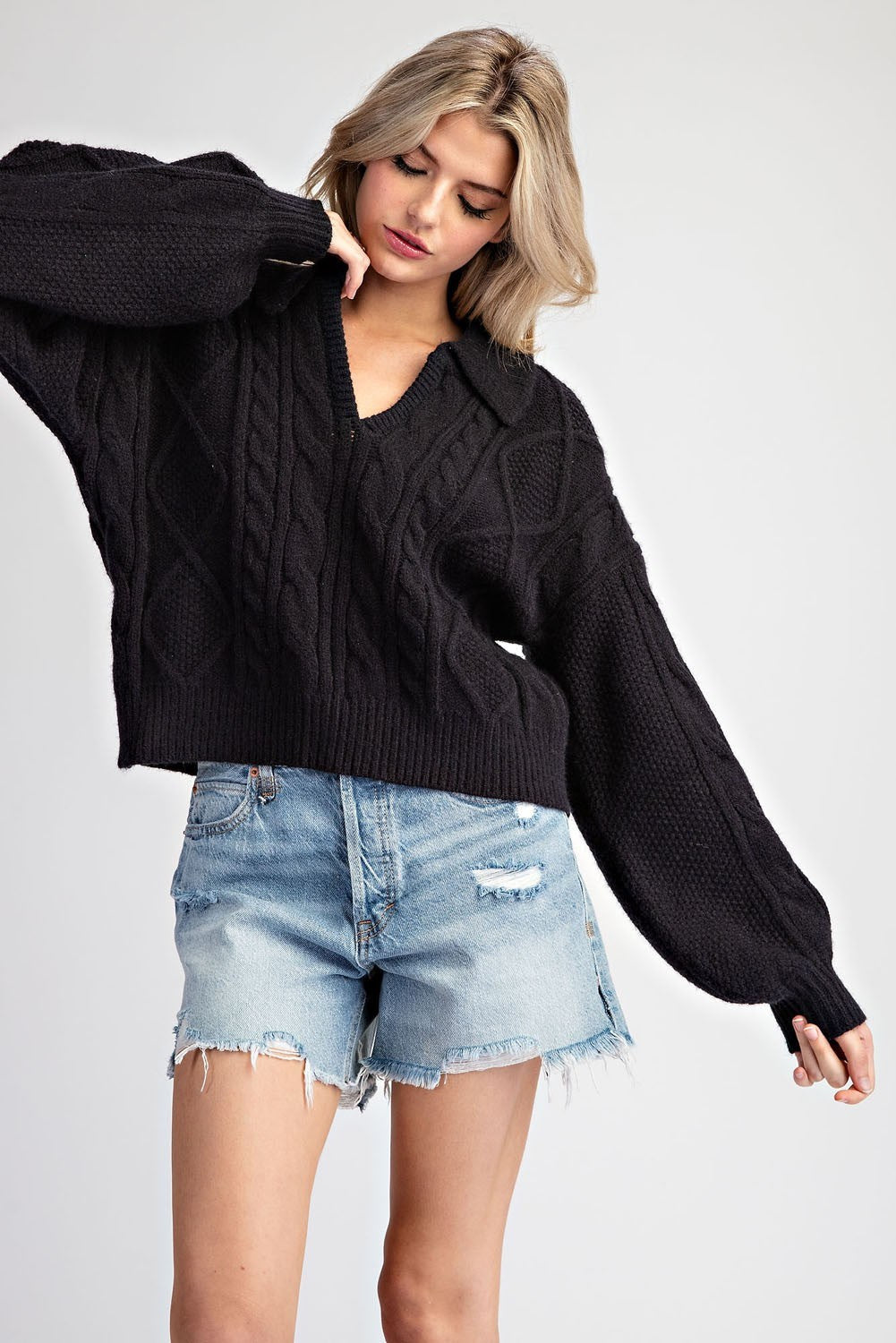 A woman wearing a black cable-knit cropped sweater with balloon sleeves and light-wash distressed denim shorts, posed against a light gray studio background.