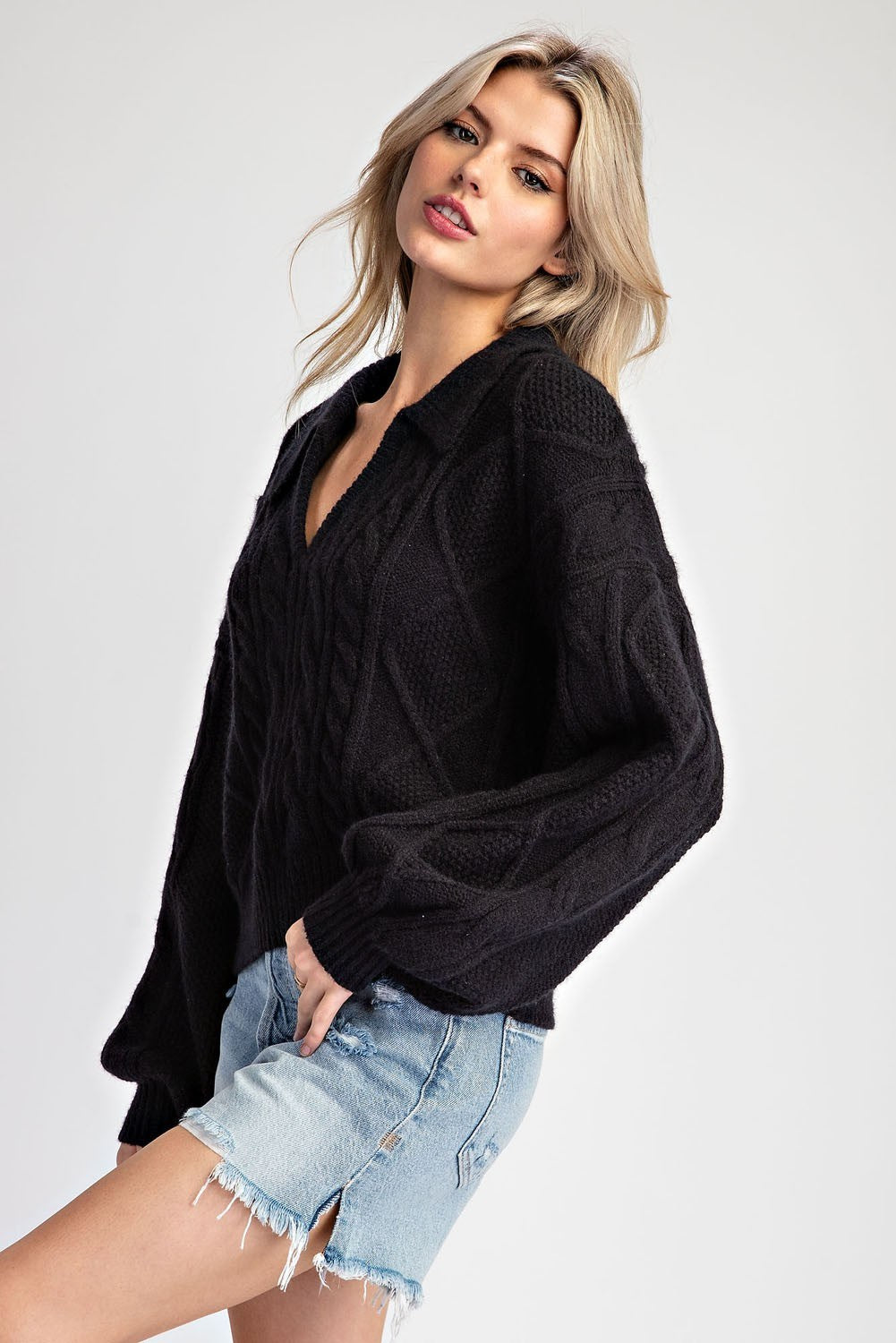 A blonde woman wearing a black cable-knit button-up sweater and light-wash denim shorts, posing against a light gray studio background.
