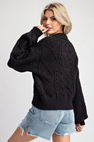 A woman wearing a black cable-knit sweater and light-wash denim shorts, photographed against a plain light gray studio background.
