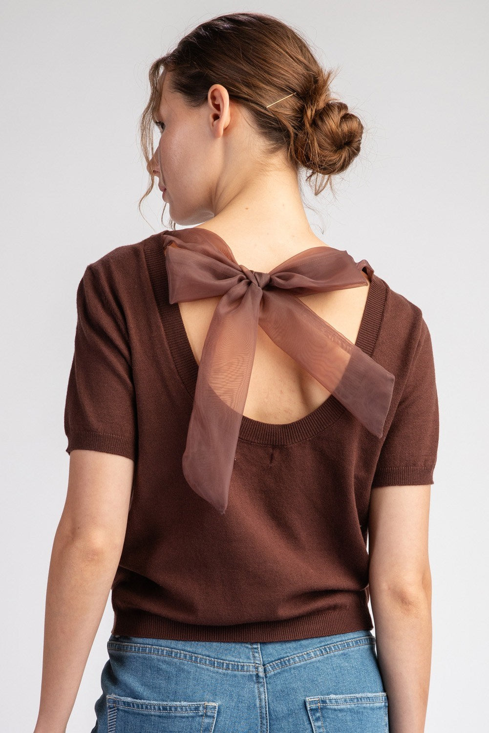 Woman wearing a brown blouse with a bow detail on the back against a plain background