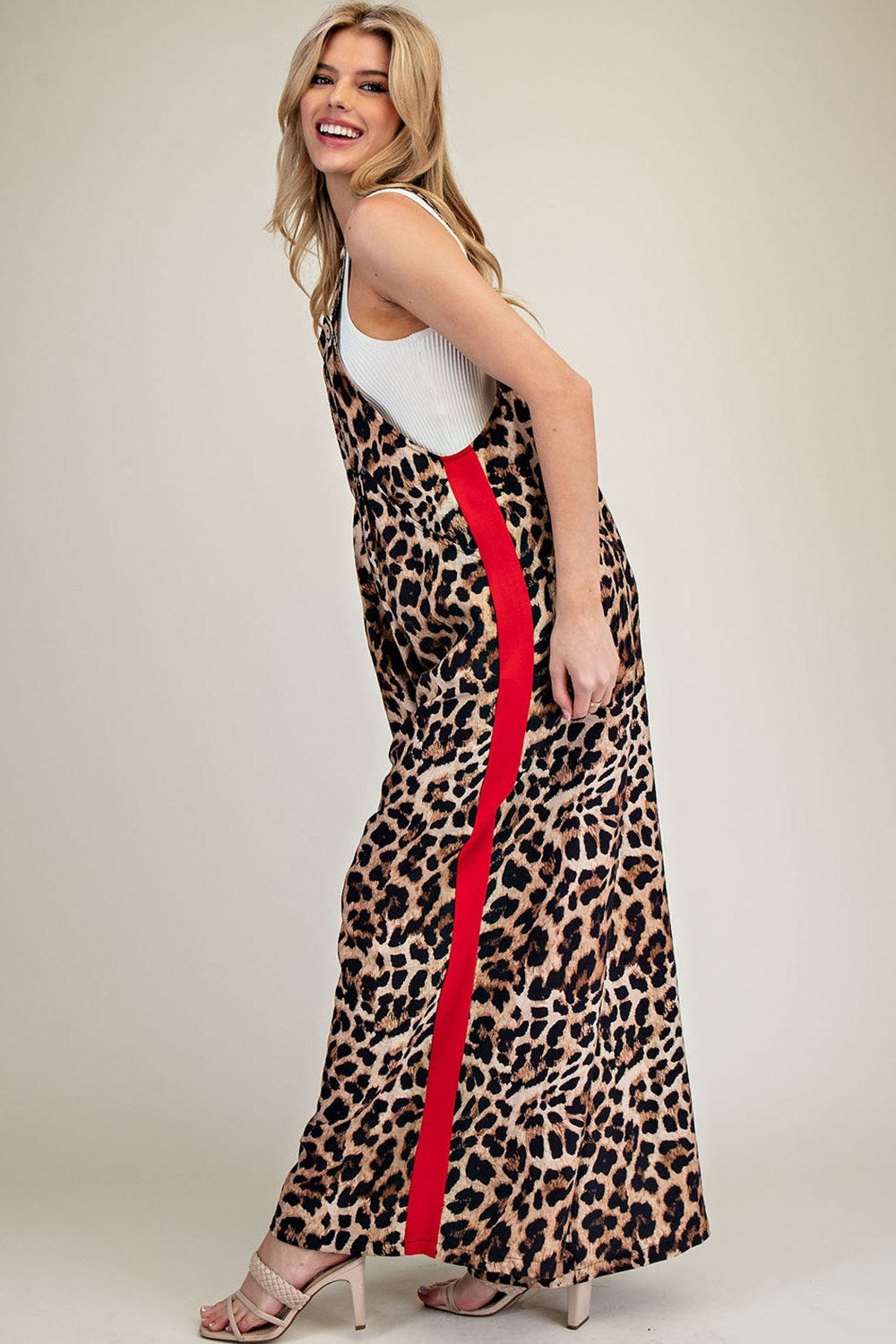 A woman modeling a sleeveless leopard-print maxi dress with a bold red side stripe, worn over a white tank top and paired with beige high-heeled sandals, set against a plain beige studio background.