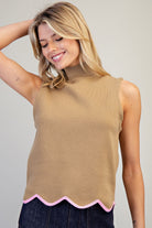 A woman wearing a sleeveless mock neck knit top in camel tan with a pink scalloped hem, paired with dark denim jeans, against a neutral light gray background.