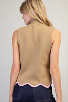 A woman shown from the back wearing a sleeveless camel-colored knit top with a pink scalloped hem, paired with dark blue jeans, against a light gray studio background.