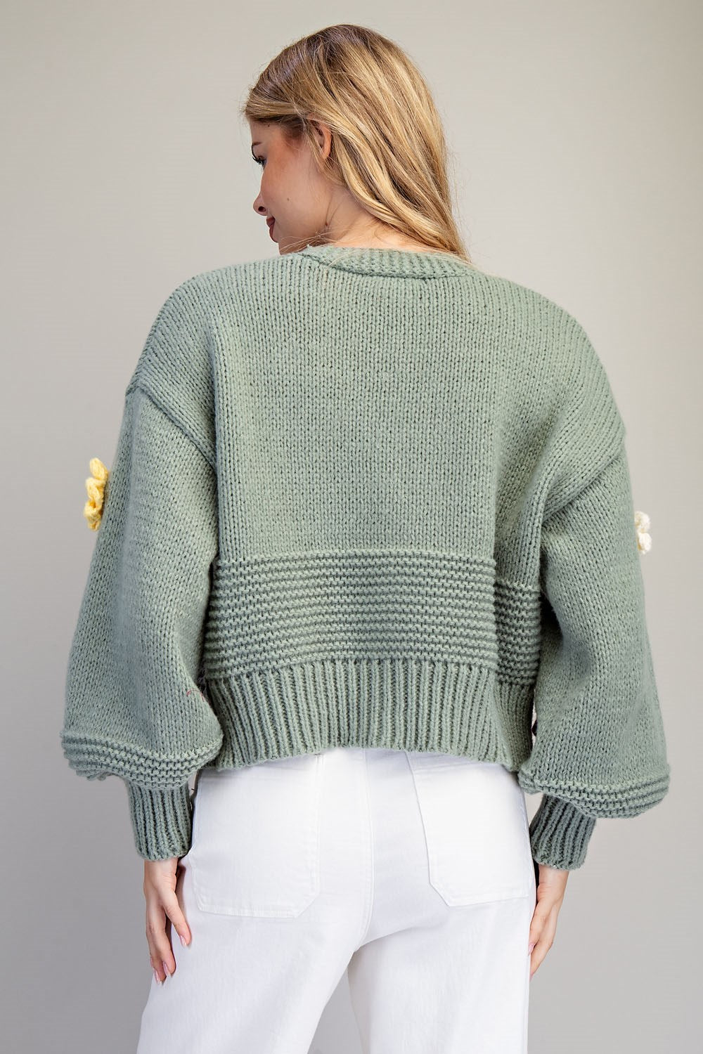 A woman shown from the back wearing a sage green chunky knit sweater with balloon sleeves and white pants, set against a plain light gray studio background.
