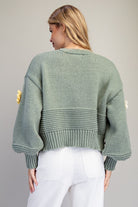 A woman shown from the back wearing a sage green chunky knit sweater with balloon sleeves and white pants, set against a plain light gray studio background.