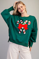 A woman wearing a dark green crewneck sweatshirt with a large sequined candy cane and red bow graphic on the front, paired with off-white corduroy pants, against a neutral gray background.