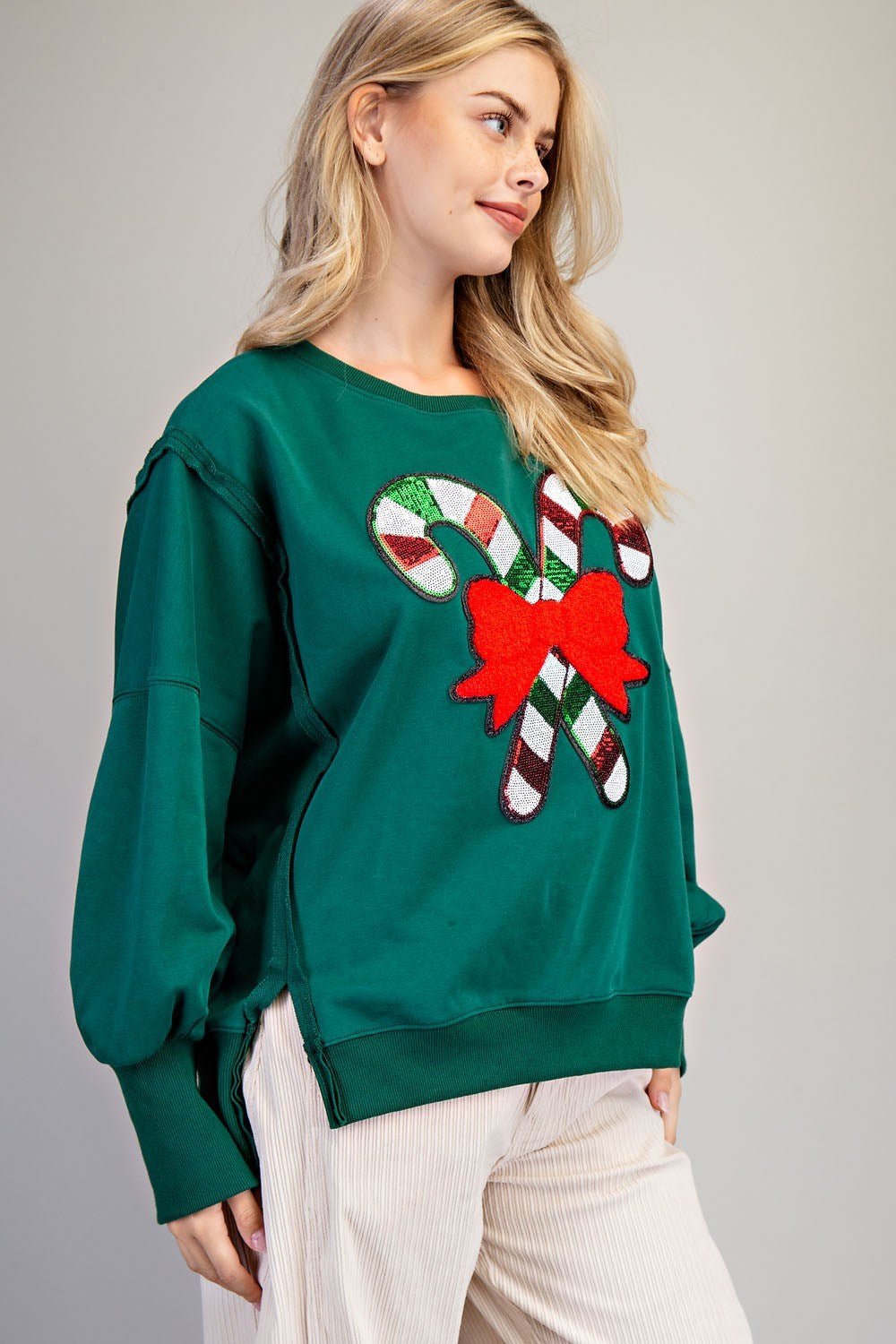 A woman wearing a dark green crewneck sweatshirt with a sequined candy cane and red bow graphic on the front, paired with off-white ribbed pants, against a neutral gray background.