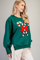 A woman wearing a dark green crewneck sweatshirt with a sequined candy cane and red bow graphic on the front, paired with off-white ribbed pants, against a neutral gray background.