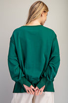 A woman viewed from behind wearing a dark green long-sleeve top with balloon sleeves and white pants, standing against a neutral gray background.