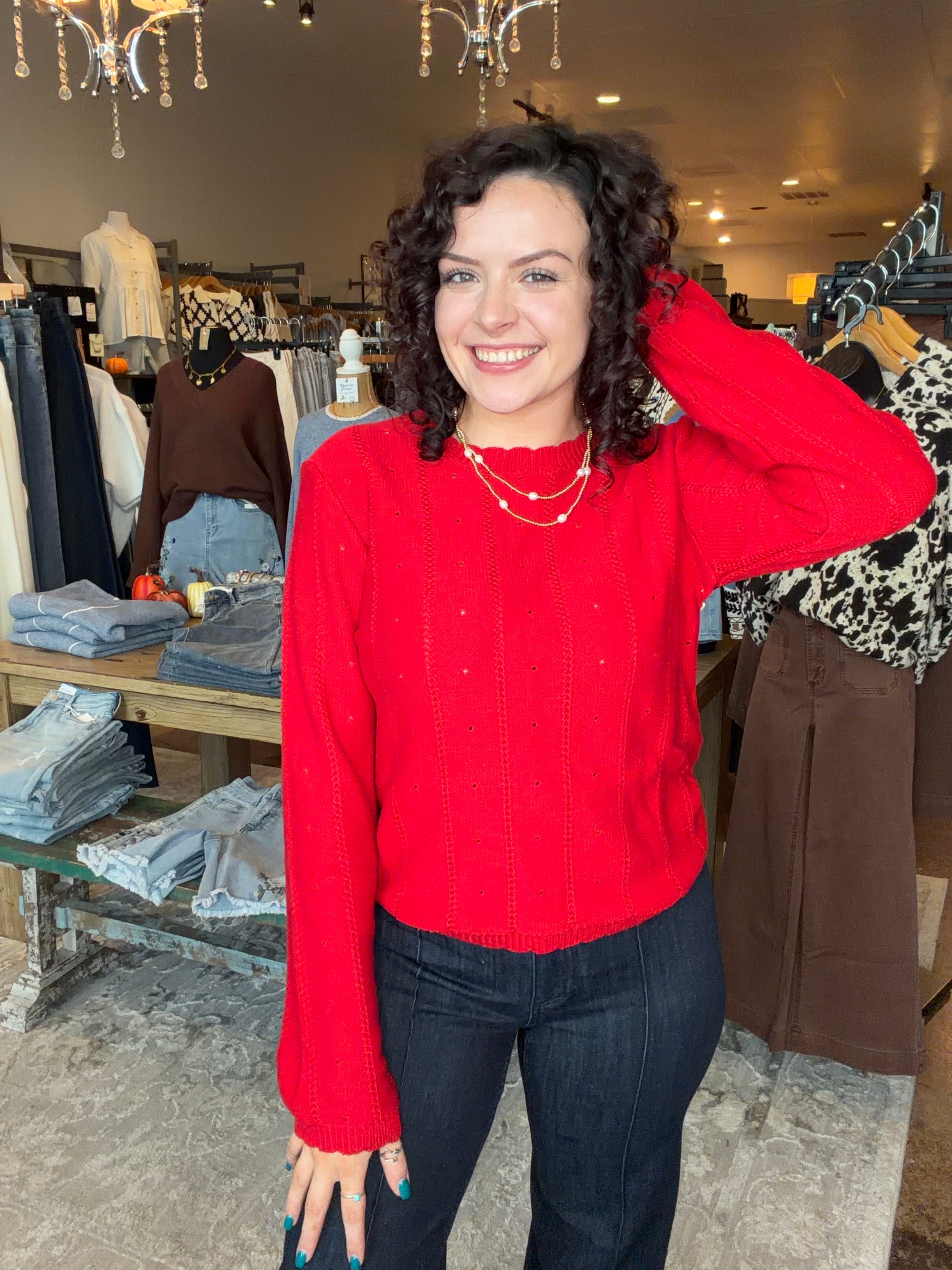 A woman with curly dark hair smiling in a clothing boutique, wearing a bright red knit sweater and dark jeans.